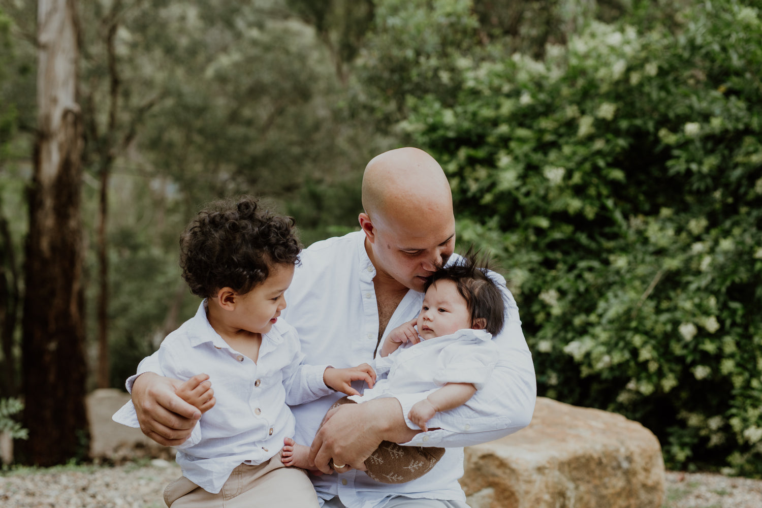 Dad holding two children outdoors with greenery in the background